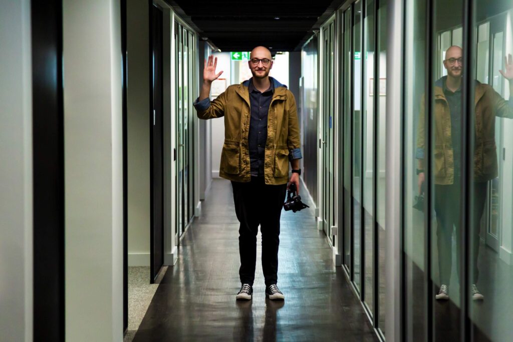 Christopher standing in a reflective hallway, with a camera in one hand and waving at the camera with the other hand