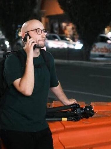 Christopher looking off to the right, holding a phone to his ear and securing a tripod with his other hand while sitting on an orange traffic bollard